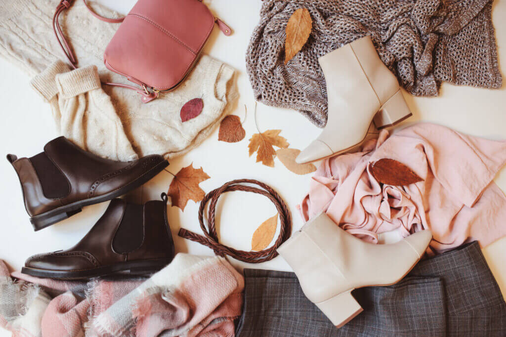 A variety of clothing and accessories is artistically laid out on a bed, with several fall leaves scattered on top.