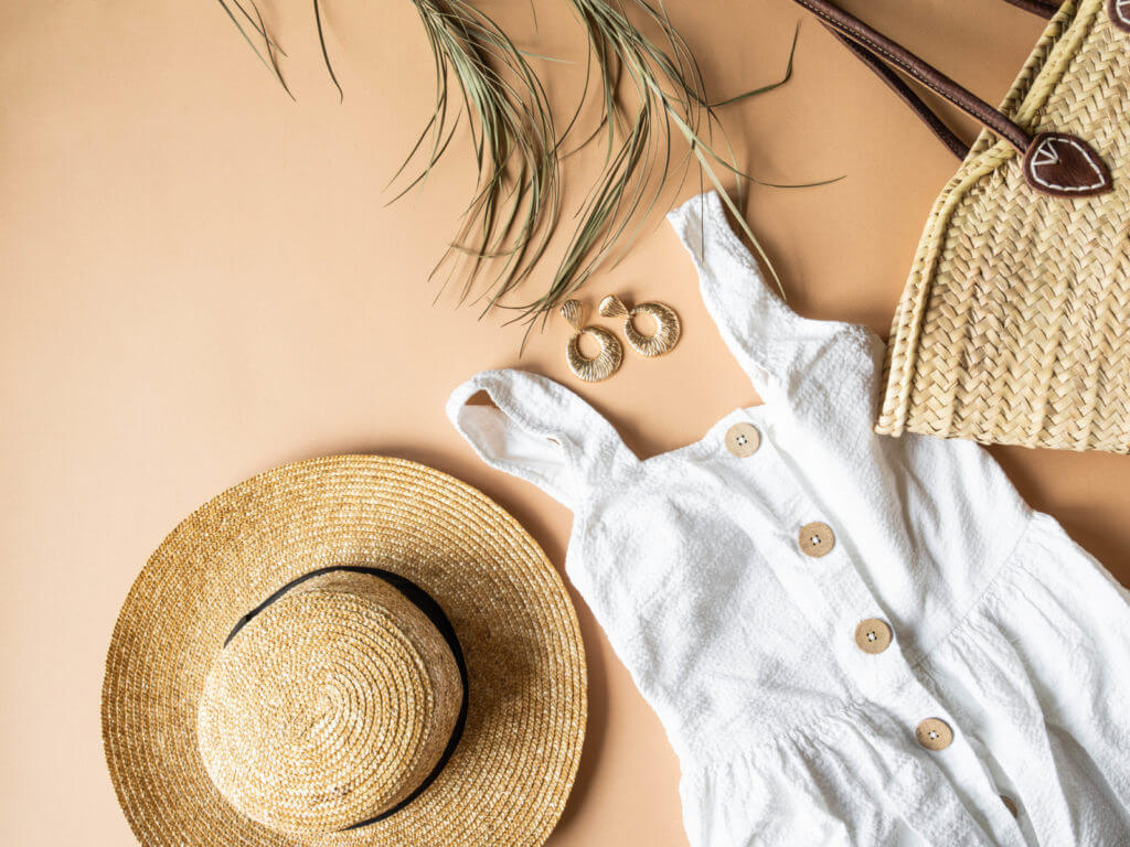A dress, and earrings are laid out on the ground next to a woven sun hat and tote bag.
