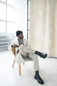 A man confidently sits in a chair wearing white pants and a white blazer.