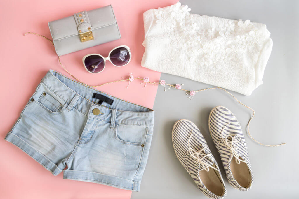 Short shorts are laid out on the ground next to cute striped tennis shoes, a flower necklace, sunglass, a wallet and a clutch with a lace floral design.